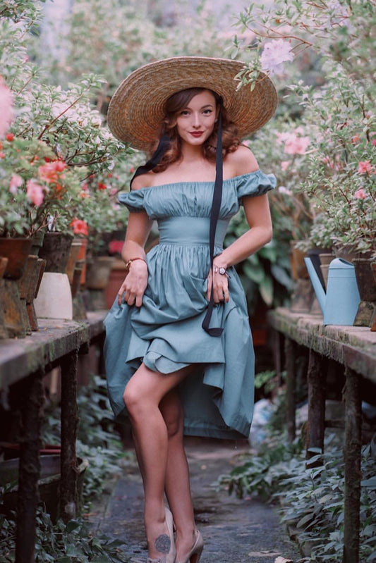 Woman in a light blue dress and straw hat standing in a garden with potted plants.